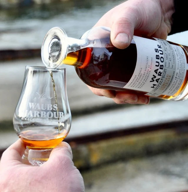 A hand holds a clear glass tumbler engraved with 'WAUBS HARBOUR' while another hand pours amber liquor from a bottle labeled 'WAUBS HARBOUR' into the glass, with a blurred outdoor waterfront background.