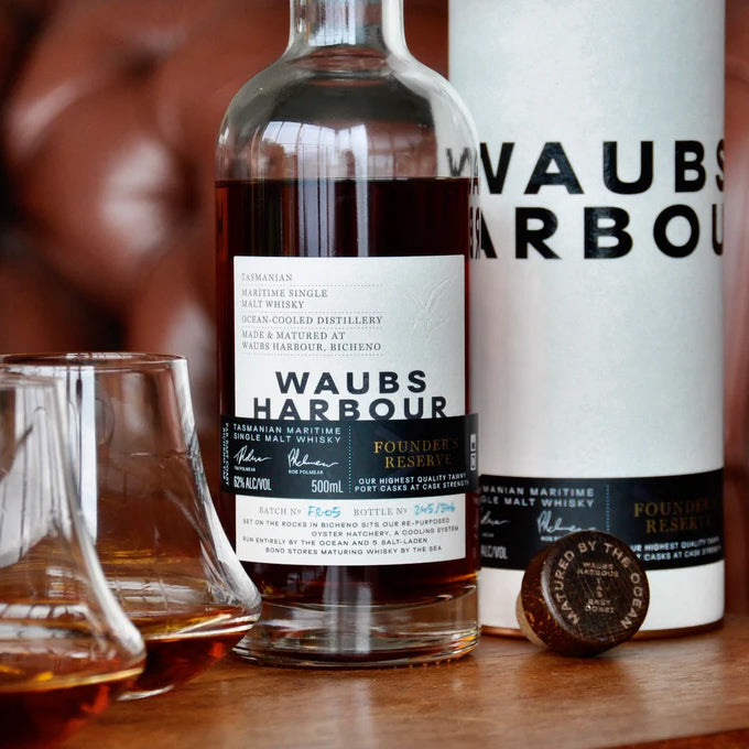 A bottle of Waubs Harbour Founders Reserve Tasmanian Single Malt Whisky is shown with its label, cylindrical box packaging, whiskey glasses filled with amber liquid, and the cap on a table—showcasing its unique maritime Tasmanian origin.