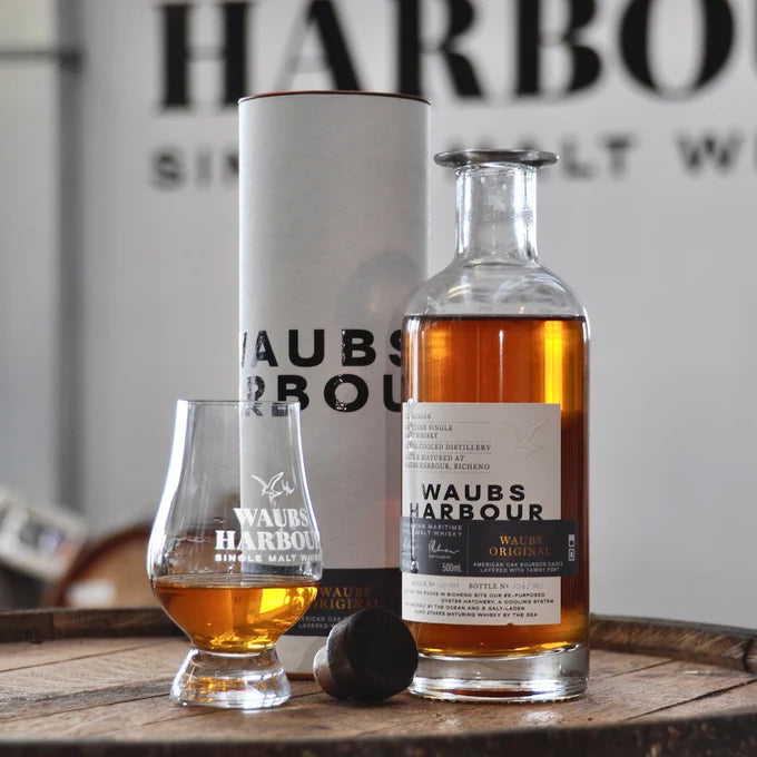 A bottle of Waubs Harbour Original Tasmanian Single Malt Whisky is displayed next to a glass, cork stopper, and its cylindrical packaging on a wooden barrel, with bold branding in the background highlighting this premium whisky experience.