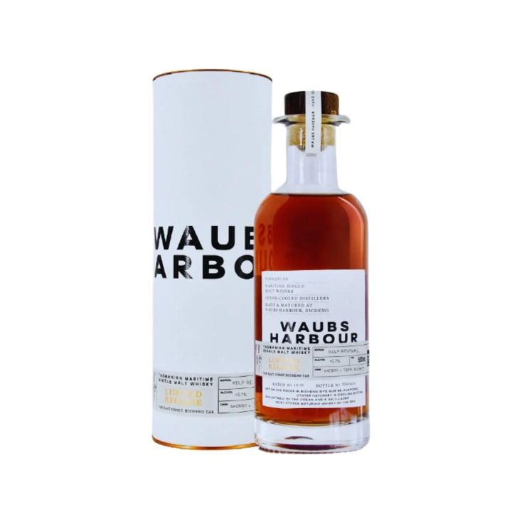 A bottle of Waubs Harbour Scotch whisky with amber liquid, next to its tall white cylindrical packaging tube, on a plain white background.