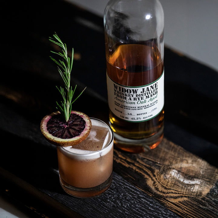 A cocktail garnished with rosemary and a slice of blood orange sits beside a bottle of Widow Jane American Oak Aged Rye Whiskey on a rustic wooden surface.