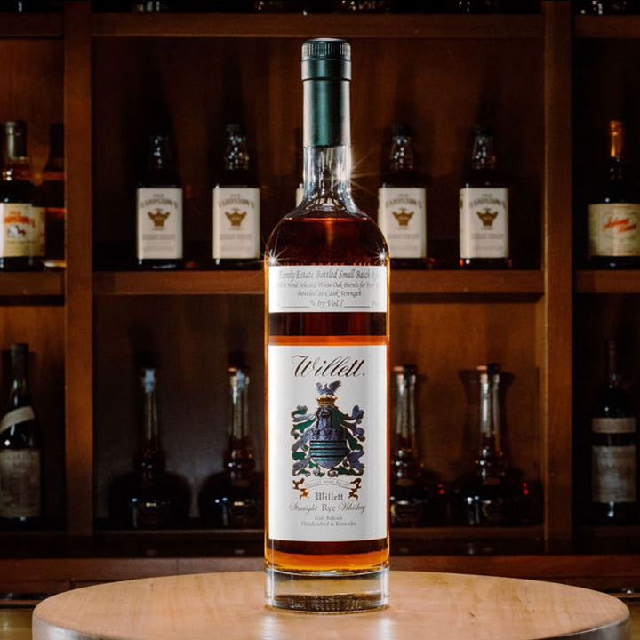 A bottle of Willett Family Estate 4 Year Old Rye Whiskey sits on a wooden surface, while shelves filled with assorted whiskey bottles appear blurred in the background.