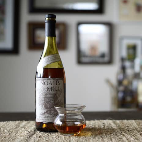 A bottle of Willett Family Estate Noah's Mill Bourbon Whiskey sits on a table beside a glass of whiskey, while blurred framed pictures decorate the wall in the background.