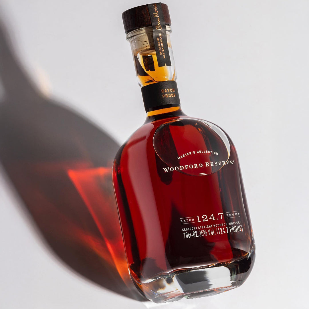 A bottle of Woodford Reserve Batch Proof 59.2% Bourbon Whiskey, 2022 edition, stands upright on a white surface with a long shadow. Amber whiskey shines through the clear glass.