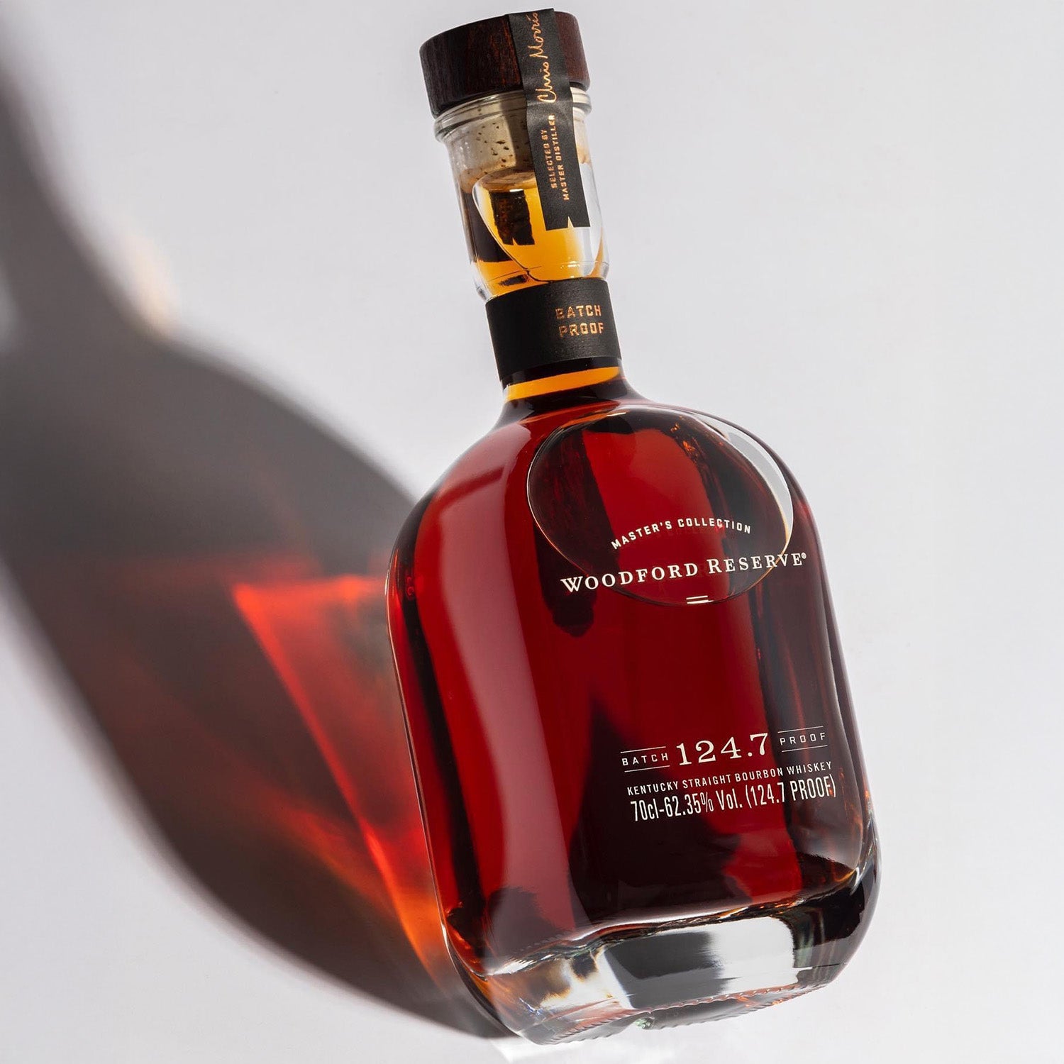 A bottle of Woodford Reserve Batch Proof 59.2% Bourbon Whiskey, 2022 edition, stands upright on a white surface with a long shadow. Amber whiskey shines through the clear glass.