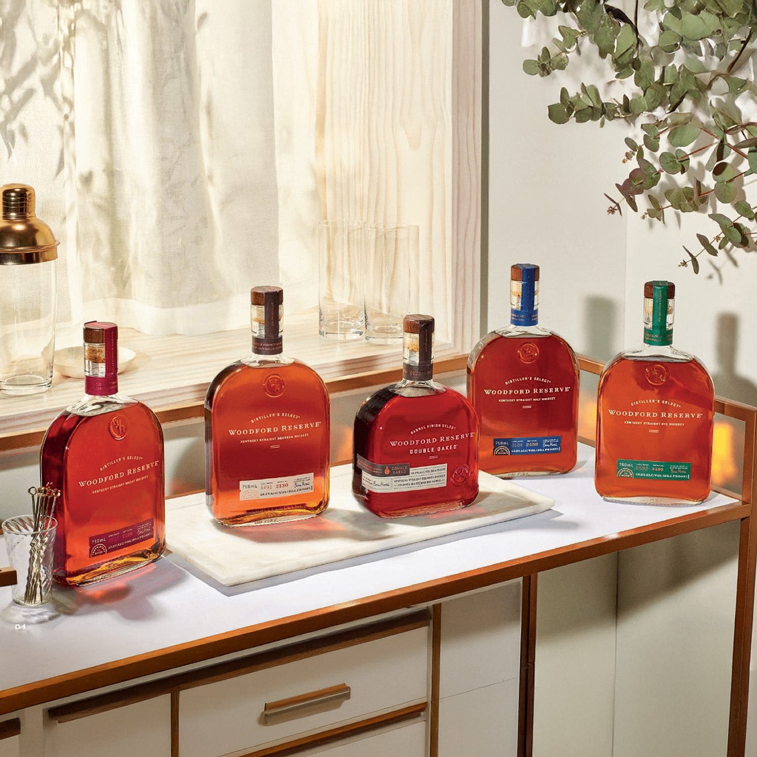 Five bottles of bourbon whiskey, including Woodford Reserve Five Malt Stout Bourbon Whiskey Master Collection, are lined up on a counter near a sunlit window with glasses and a cocktail shaker nearby.