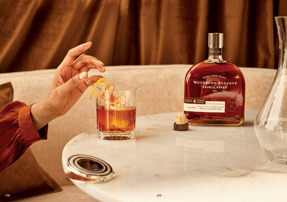 A hand garnishes a glass of bourbon with an orange twist on marble. Beside the glass is a bottle of Woodford Reserve Double Oaked Bourbon Whiskey, known for its double-barrel aging, along with a stopper and a nearby decanter.