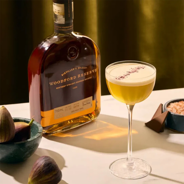 A bottle of Woodford Reserve Kentucky Straight Bourbon Whiskey stands beside a coupe glass holding a frothy cocktail with an herb garnish, surrounded by fresh figs, chocolate, and a small bowl of pink salt on a light surface.