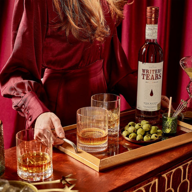 Person pouring Writer's Tears whiskey into glasses on a wooden tray with a red curtain background.