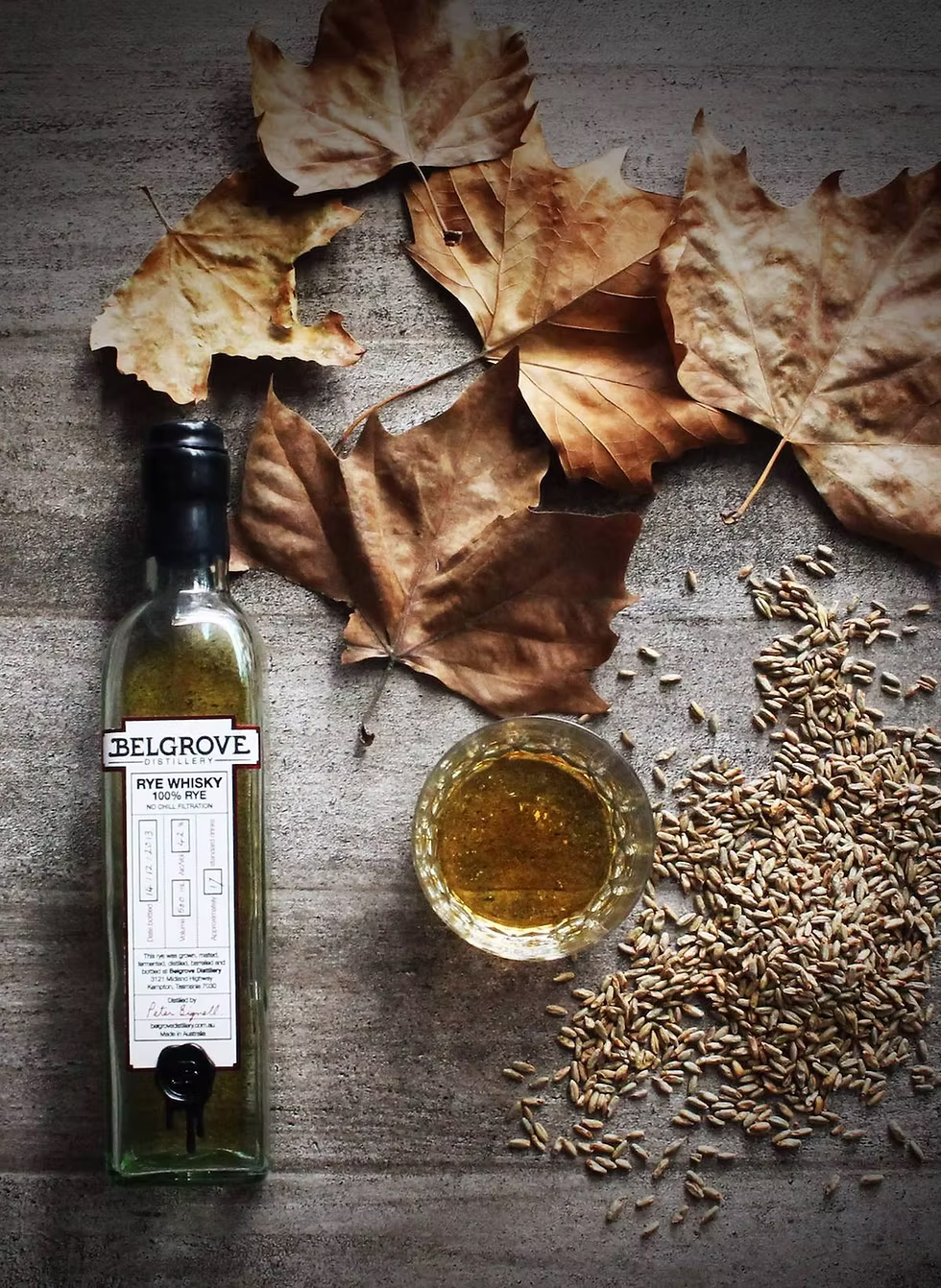 Belgrove whiskey bottle, glass, and seeds on a wooden surface with autumn leaves.