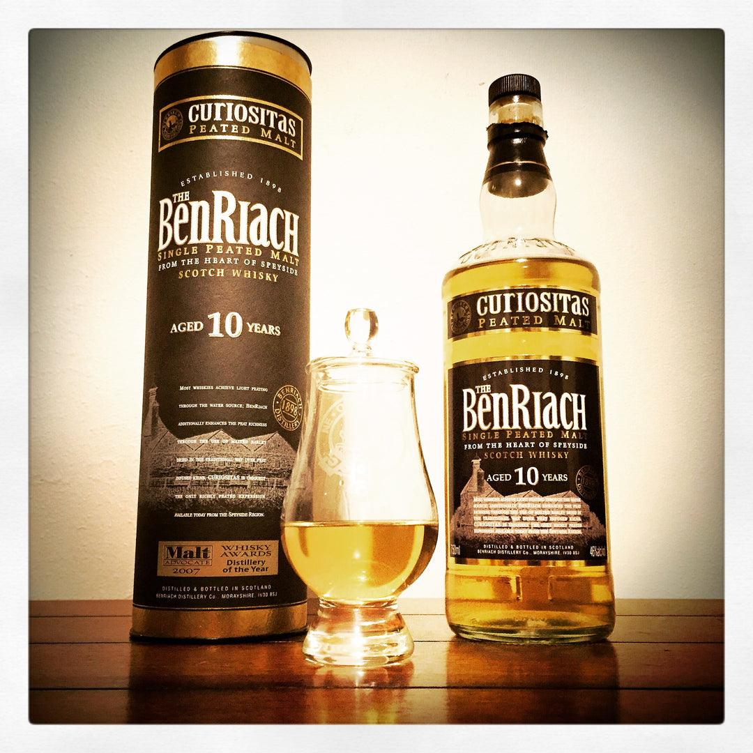 A bottle of Cutiositas Ben Riach aged 10 years single malt Scotch whisky, shown next to its matching cylindrical tin packaging and a small tulip glass filled with the golden whisky, set on a wooden surface against a plain background.