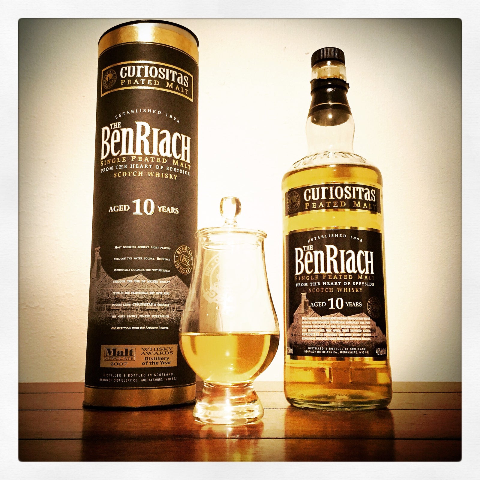 A bottle of Cutiositas Ben Riach aged 10 years single malt Scotch whisky, shown next to its matching cylindrical tin packaging and a small tulip glass filled with the golden whisky, set on a wooden surface against a plain background.