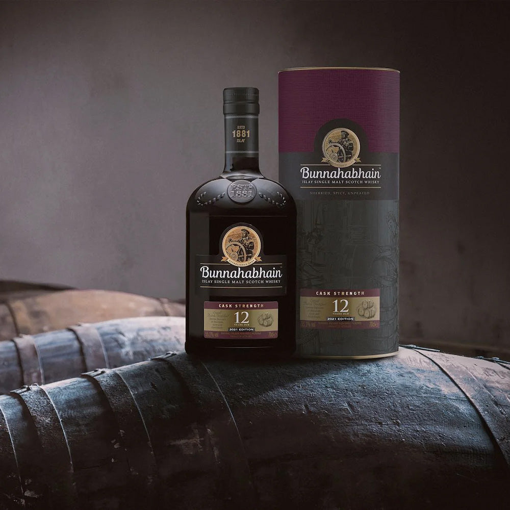 A bottle and cylindrical box of Bunnahabhain 12 Year Old Cask Strength Single Malt Scotch Whisky rest on a wooden barrel, with more barrels and a dimly lit background behind them.