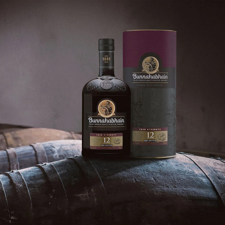 A bottle and cylindrical box of Bunnahabhain 12 Year Old Cask Strength Single Malt Scotch Whisky rest on a wooden barrel, with more barrels and a dimly lit background behind them.