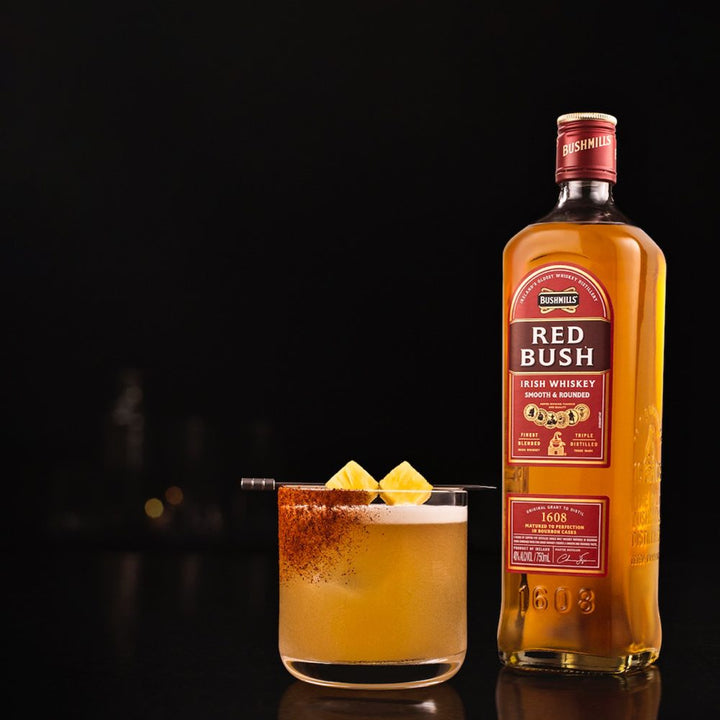 Bushmills Red Bush Irish Whiskey, matured in bourbon casks, is displayed beside a cocktail topped with pineapple chunks and a spiced rim of cinnamon or chili powder, all set against a dark background.