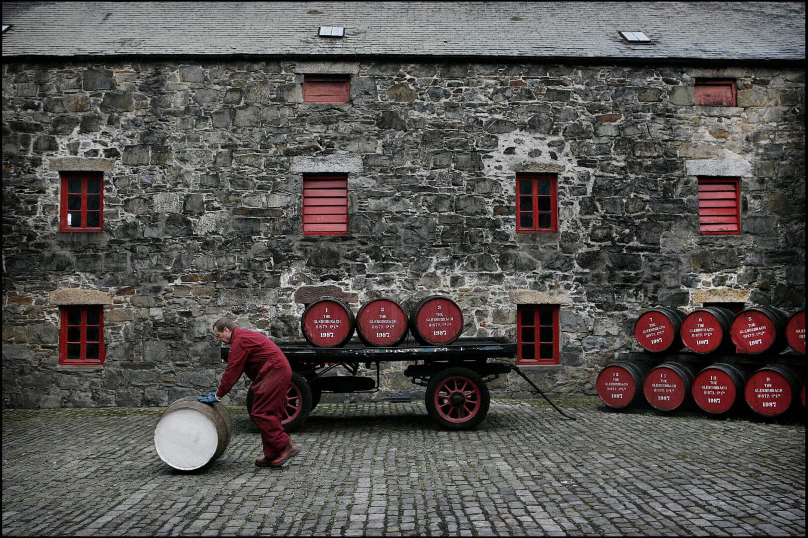 Person in red overalls pushing a cart with barrels in front of a stone building with red windows.