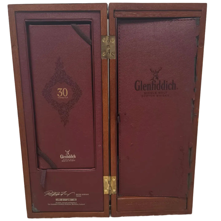 A Glenfiddich 30-year-old single malt Scotch whisky box, opened to show the dark reddish-brown interior packaging, standing on a beige carpeted floor against a white paneled wall.