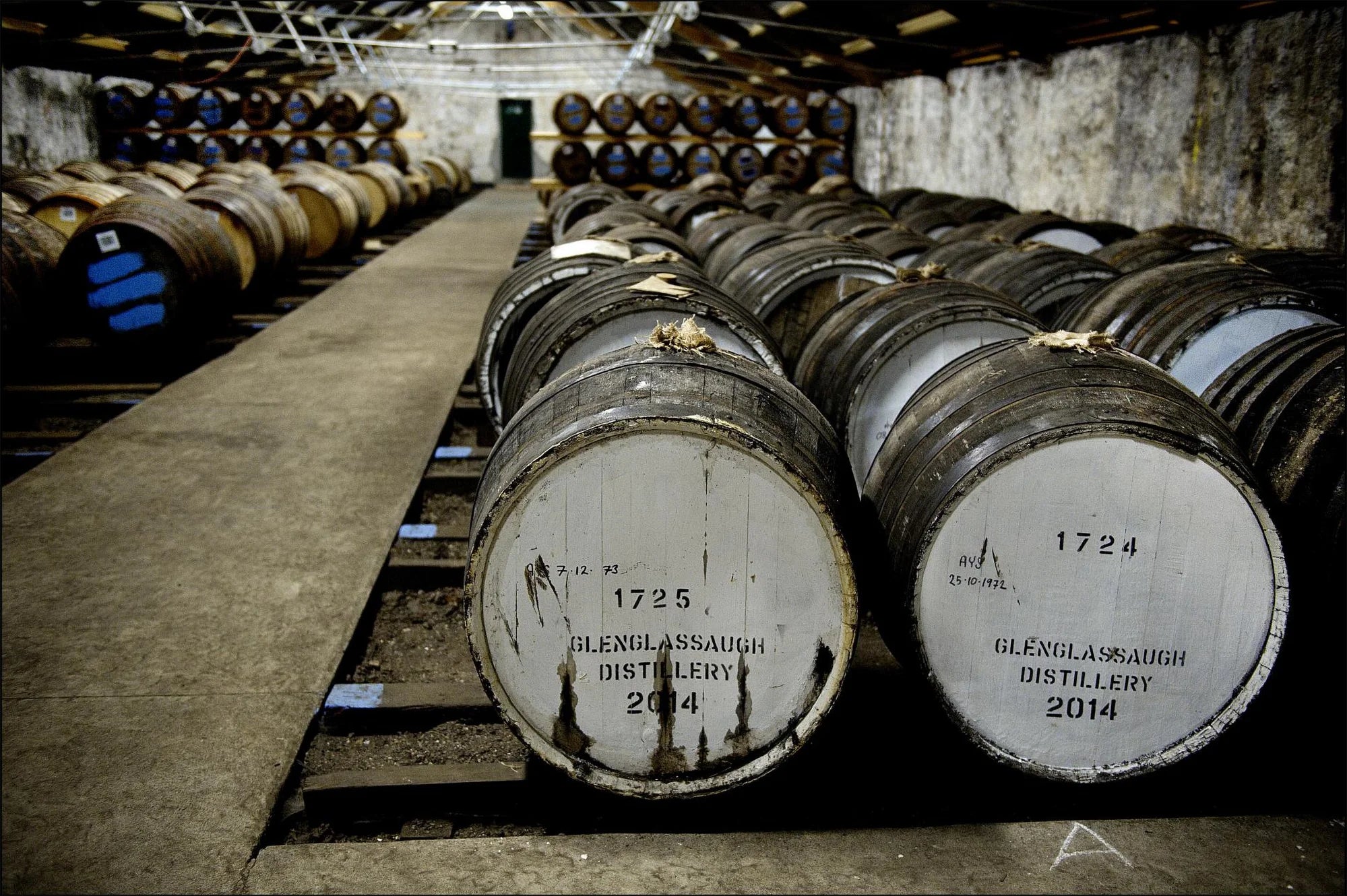 Row of whisky barrels with 'GlenGlassaugh Distillery' labels in a warehouse.