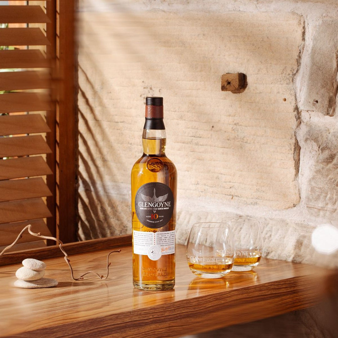 A bottle of Glengoyne 10 Year Old Single Malt Scotch Whisky sits on a wooden table beside two partly filled glasses, as sunlight streams through blinds onto a stone wall, showcasing this smooth, unpeated Scotch.