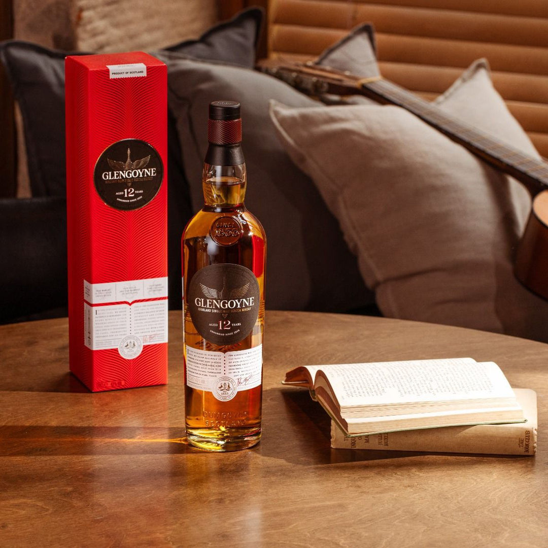 A bottle of Glengoyne 12 Year Old Single Malt Scotch Whisky stands next to its red box on a wooden table, with an open book and a couch with pillows in the background.