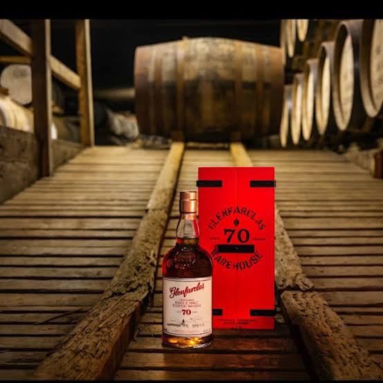 A bottle of Blachford 70 Year Old single malt whisky stands next to its red packaging box on a wooden plank pathway inside a warehouse, with wooden barrels blurred in the background.
