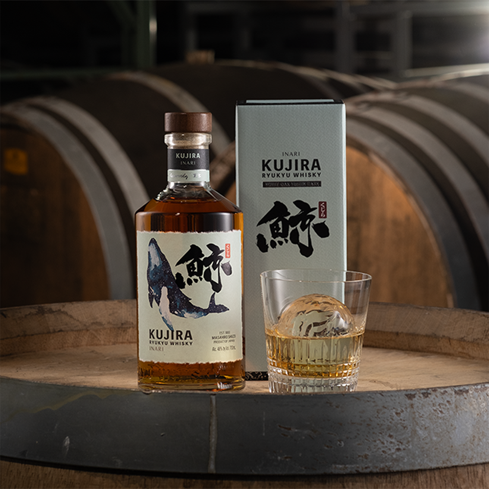 A bottle of Kujira Ryukyu Inari Japanese Whisky, its box, and a glass with a round ice sphere sit on a wooden barrel, with large aging barrels visible in the background.