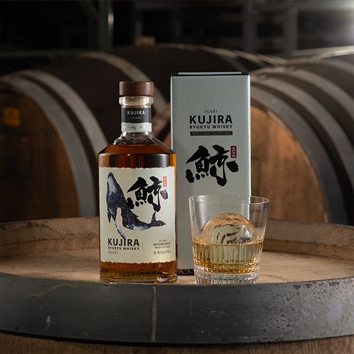 A bottle of Kujira Ryukyu Inari Japanese Whisky, its box, and a glass with a round ice sphere sit on a wooden barrel, with large aging barrels visible in the background.