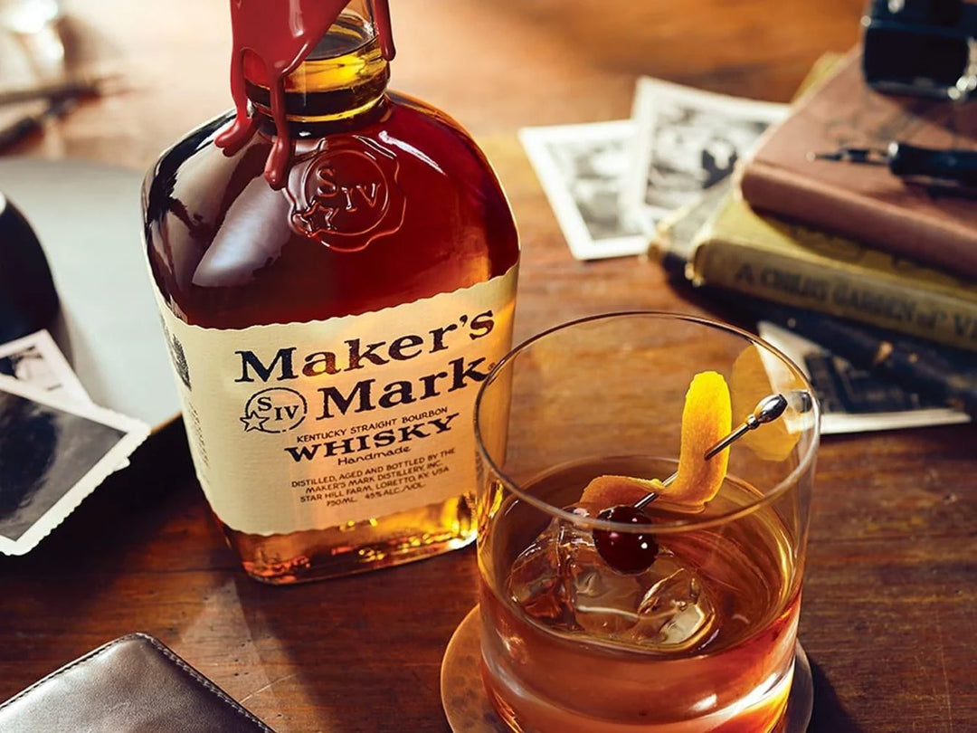 A bottle of Maker's Mark 14 Year Old Whisky stands on a wooden table beside a rocks glass filled with amber liquor and an orange peel garnish, with blurred books and papers in the background.