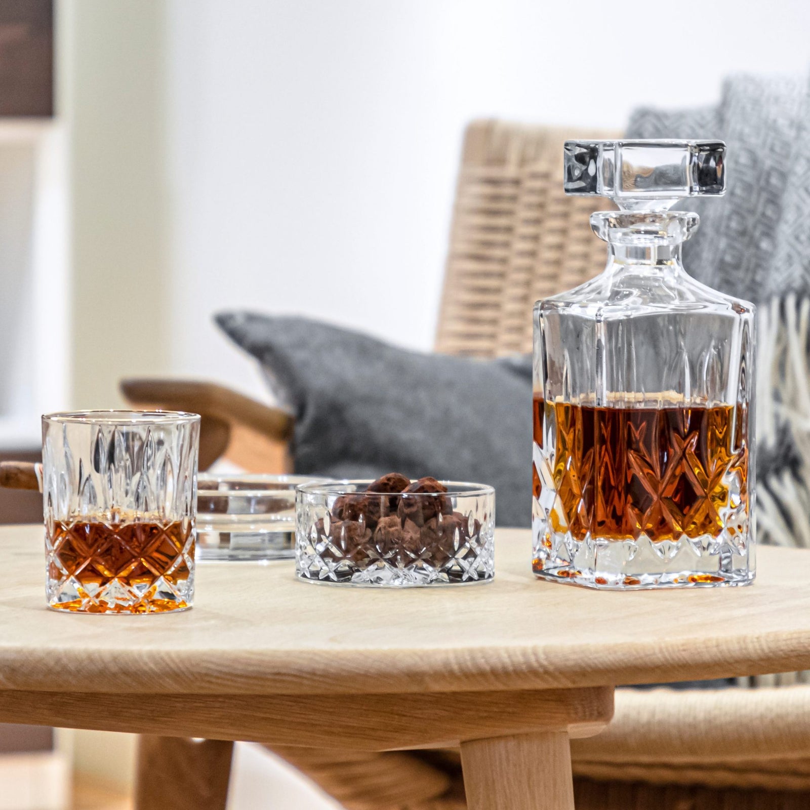 A Nachtmann Noblesse Whisky Decanter Set, featuring a crystal decanter and glass filled with amber whisky, sits on a wooden table beside chocolate truffles, with a woven chair and cushions softly blurred in the background.