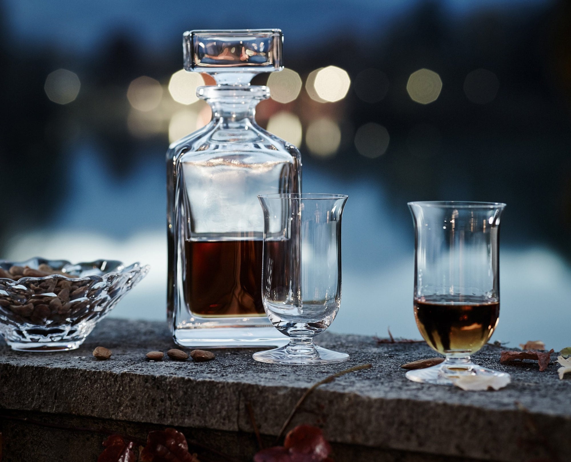Glass decanter and two glasses with a dark liquid on a stone surface, blurred lights in the background