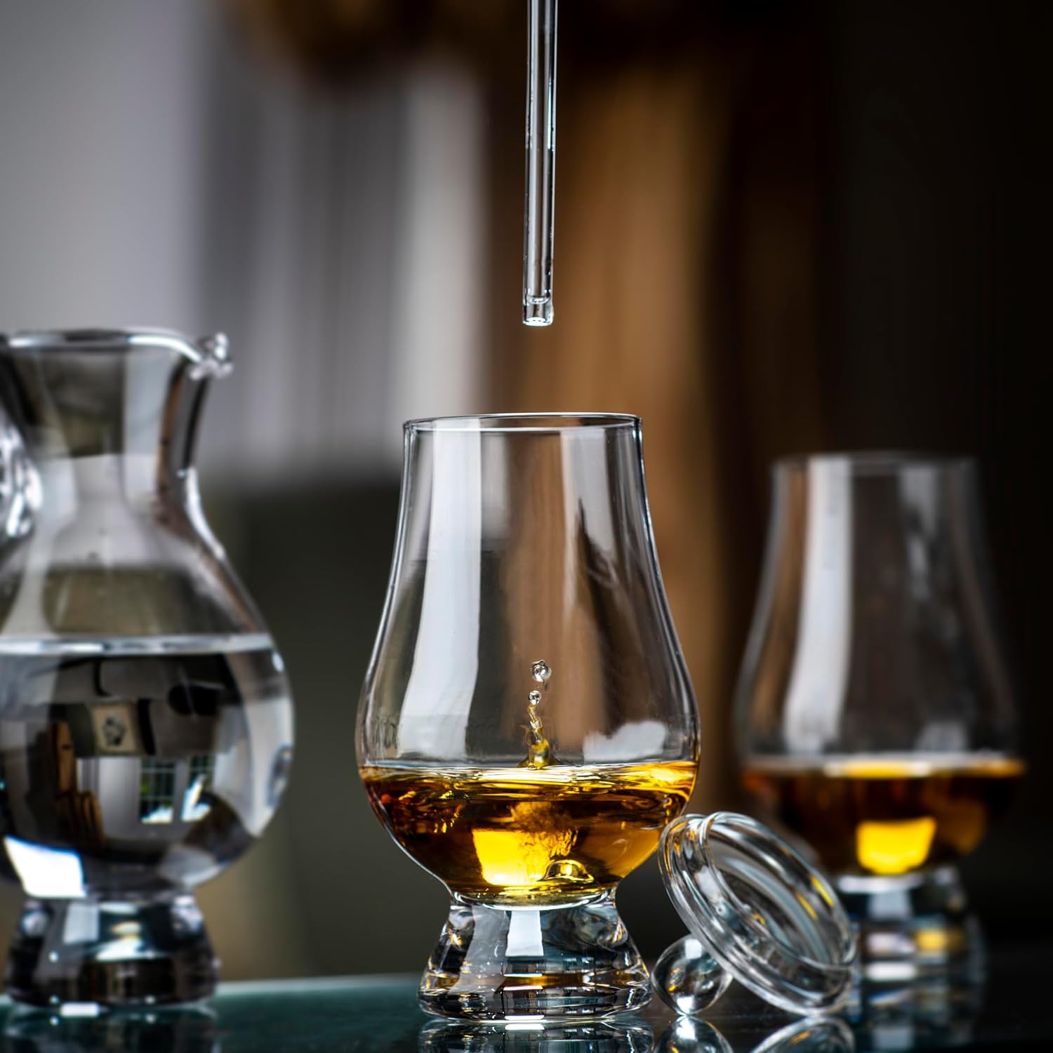 A close-up shows a whisky glass with amber liquid and a falling drop, with the Glencairn Crystal Water Jug and another glass in the background. A glass lid rests beside the front glass—ideal for whisky tasting.
