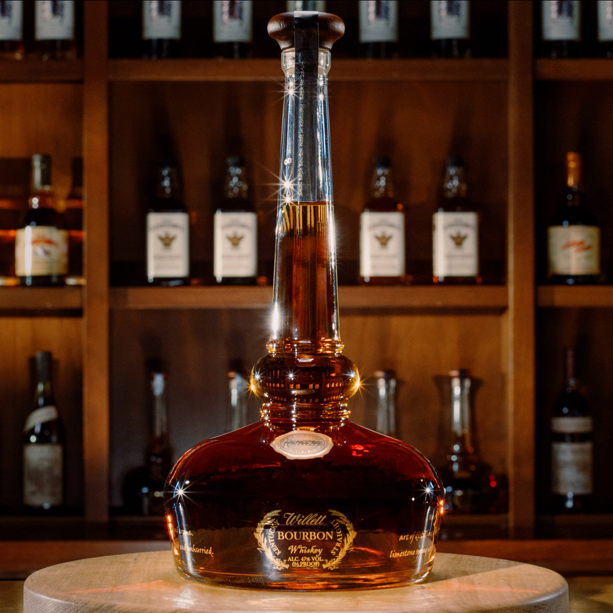 A large bottle of Willett Family Pot Still Reserve Bourbon Whiskey with a tall neck and rounded base sits on a wooden table, its amber liquid highlighted by warm lighting. Blurred shelves of whiskey bottles appear in the background.