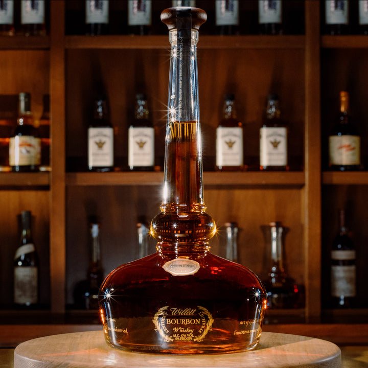 A large bottle of Willett Family Pot Still Reserve Bourbon Whiskey with a tall neck and rounded base sits on a wooden table, its amber liquid highlighted by warm lighting. Blurred shelves of whiskey bottles appear in the background.