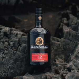 Scotch favourite, Bunnahabhain 12 year old sitting amongst black rocks to match it's dark glass bottle. A smooth and enjoyanble dram - Whisky Estate