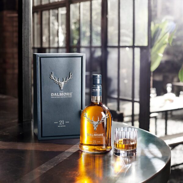 The bottle, presentation box and a glass of neat Dalmore 21 Year Old Singel Malt Whisky from Scotland atop a dark time table beside an open window with sunshine pouring inside - Whisky Estate
