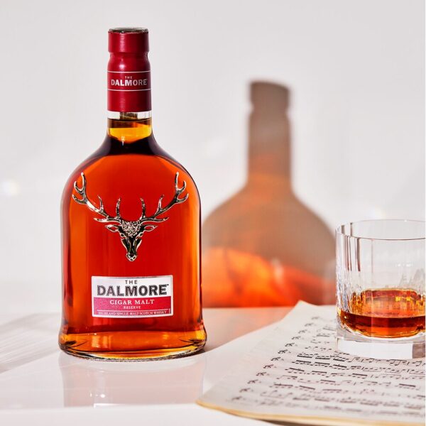 An open bottle of Dalmore Cigar Malt Reserve sits beside a glass of neat whisky, its rich amber liquid glowing. Sheets of music lie beside the bottle, adding an elegant touch to the scene, evoking a sense of sophistication and relaxation. - Whisky Estate