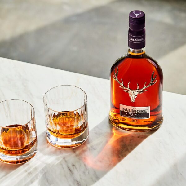 A bottle of Dalmore Port Wood Reserve rests on a marble tabletop, accompanied by two glasses of neat whisky, showcasing its deep amber colour and refined presentation. - Whisky Estate