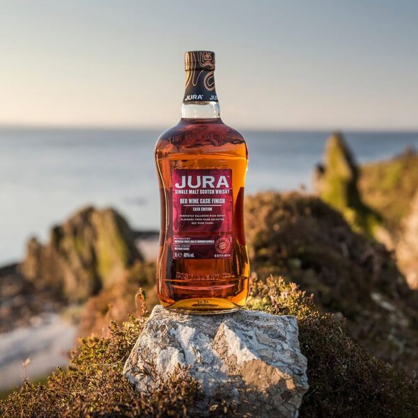 Jura Red Wine Cask Finish Single Malt whisky bottle perched atop a rock with an island landscape view, this whisky offers a smooth balance of berries, vanilla, and oak, with ex-bourbon and red wine cask maturation enhancing its fruity character. - Whisky Estate